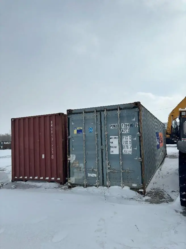 Two shipping containers on a snowy field with a part of heavy machinery visible on the right.